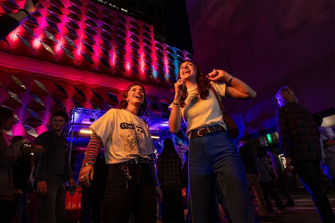 Two women smile and dance beneath a geometric ceiling glowing red and purple in a crowded nightclub, creating a lively, joyful atmosphere.