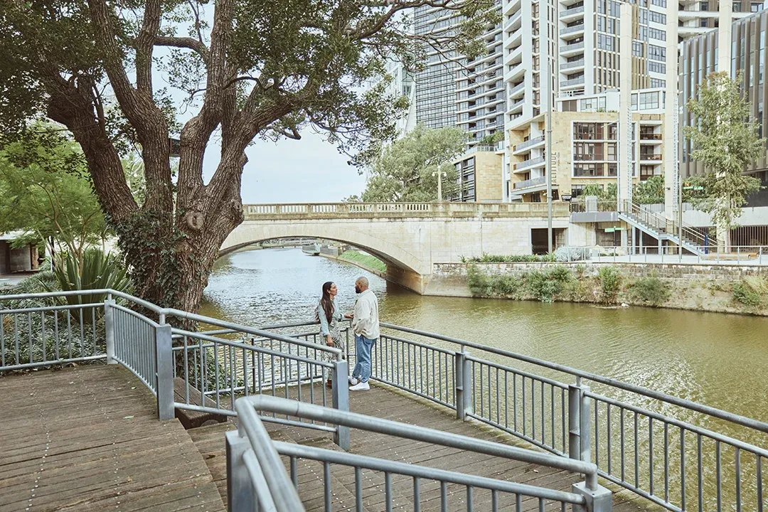A couple stands on a riverside boardwalk beneath a sprawling tree, overlooking a stone bridge and modern apartment towers in a calm urban setting.