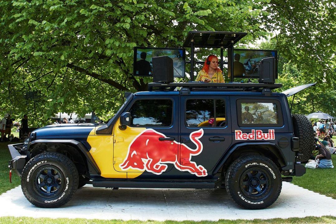 A black-and-yellow Red Bull Jeep with bull graphics and roof-mounted screens is parked under leafy trees; a woman appears on the central screen.