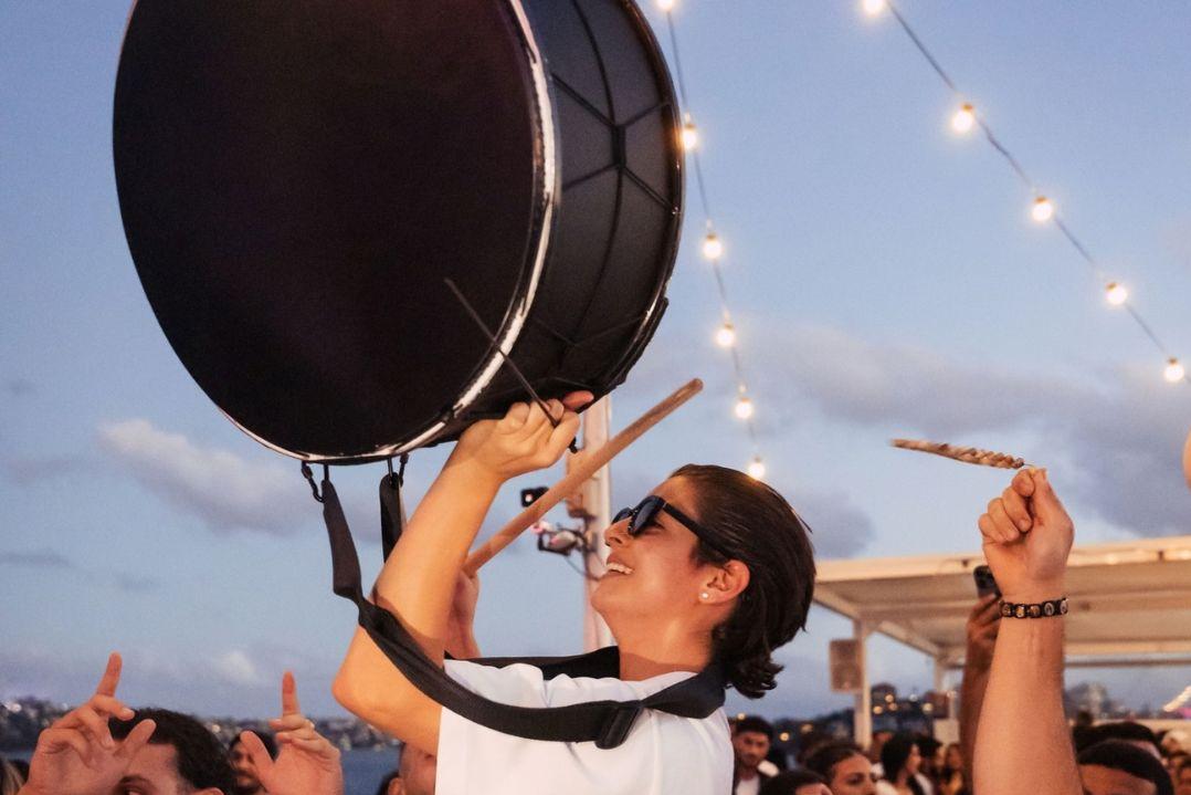 A smiling woman in sunglasses raises a large black drumstick overhead at an outdoor evening concert, as string lights glow above the cheering crowd.