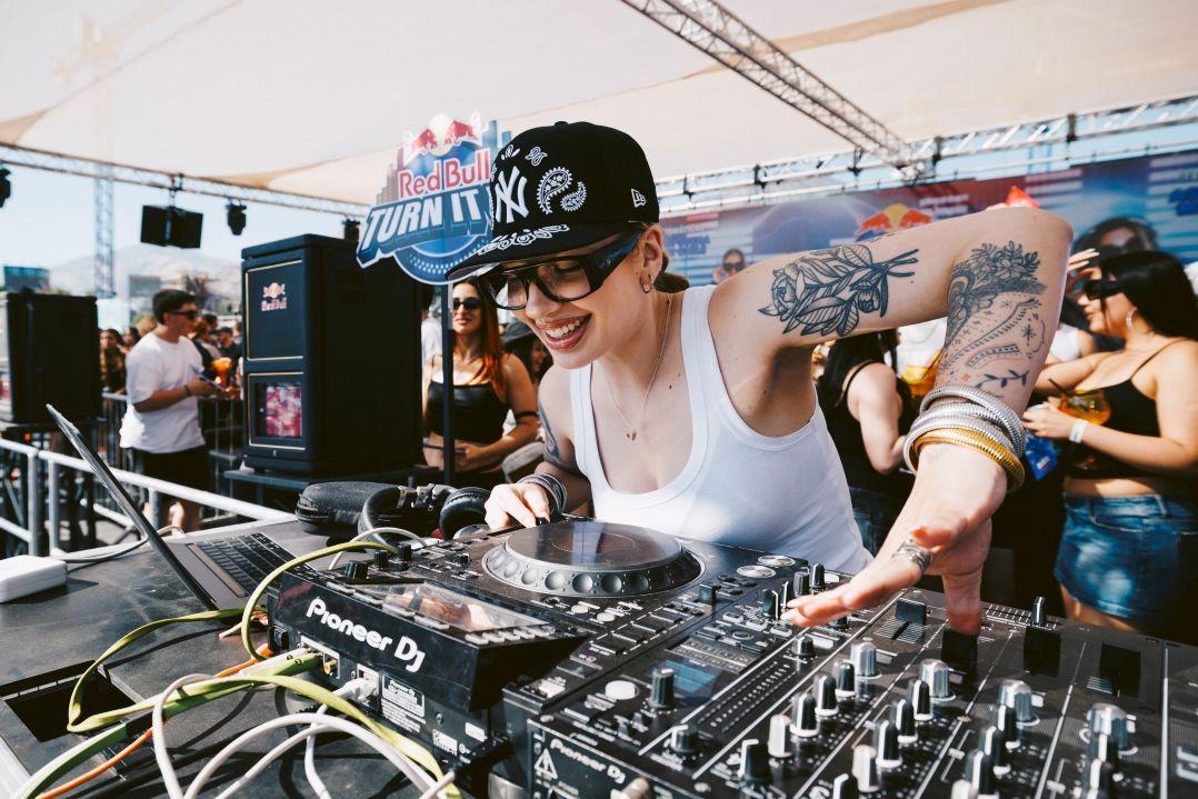A smiling, tattooed DJ in a black cap and white tank top mixes Pioneer decks at a sunny Red Bull event, with a lively crowd behind her.