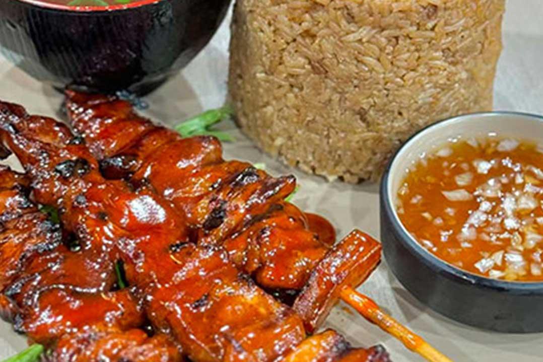 Glazed, char-grilled meat skewers fill the foreground beside a bowl of orange dipping sauce and a container of rice, creating a hearty meal.