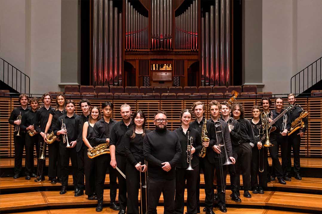 A large student orchestra, dressed in black and holding brass and woodwind instruments, poses on concert hall steps before a towering pipe organ; formal, celebratory mood.