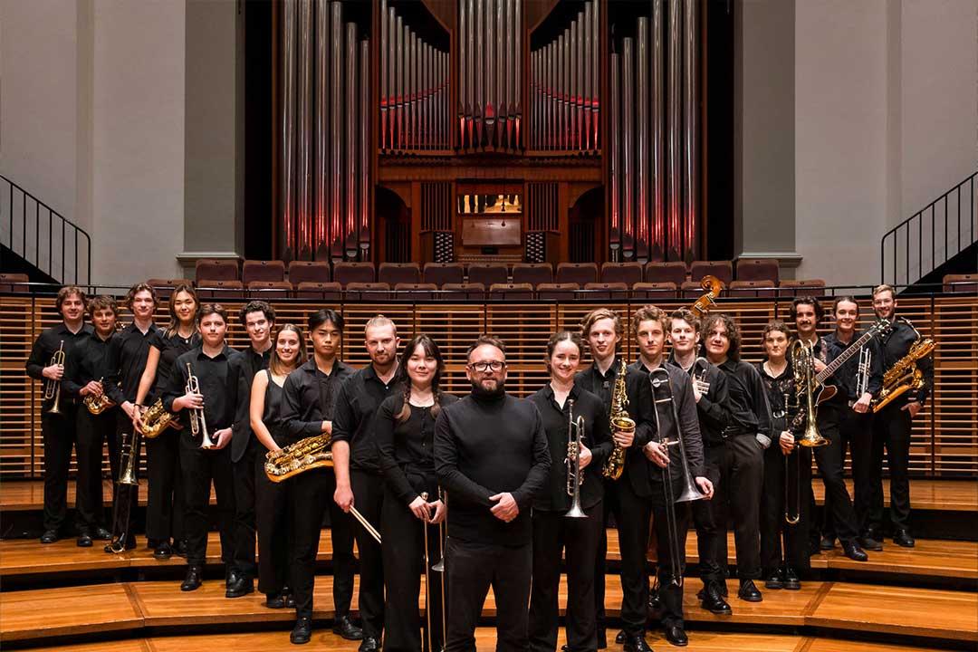 A large orchestra ensemble in black formal clothing poses with brass, woodwind, and percussion instruments on concert hall steps beneath a towering pipe organ; the mood is formal and celebratory.