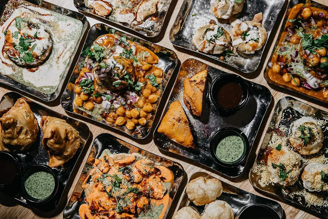 Overhead Indian feast in metal trays: chickpea curry, grilled and creamy dishes, breads, dumplings, and dipping sauces create a rich, festive spread.