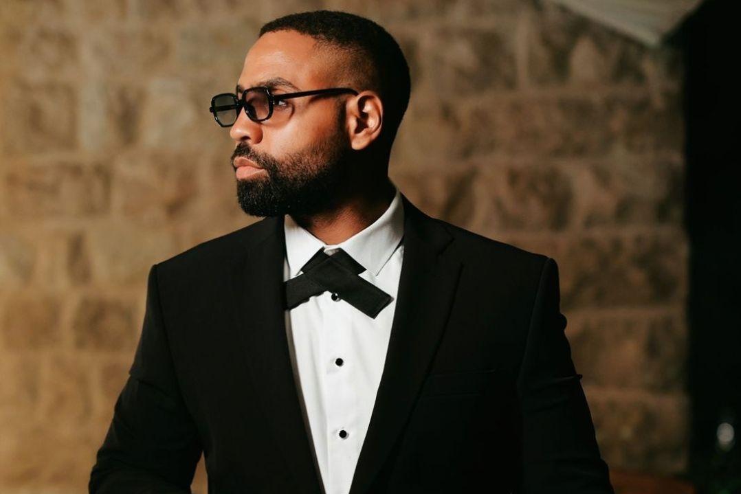 A bearded man in black glasses and a tuxedo stands against a warm stone wall, looking thoughtfully to the side with a poised, formal air.