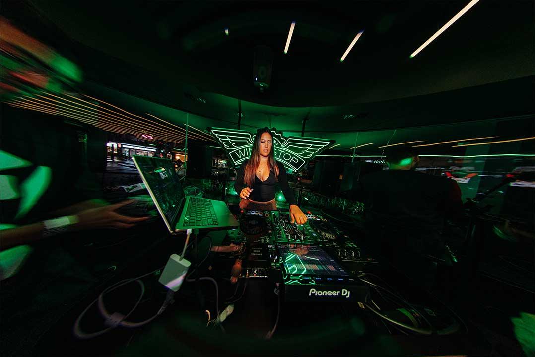 A female DJ performs behind Pioneer DJ decks in a dark nightclub, lit by vivid green lasers and neon accents, creating an energetic, immersive mood.