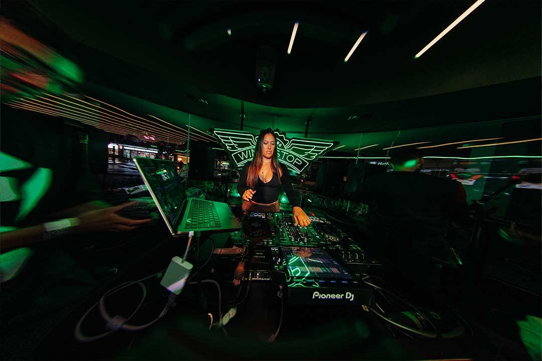 A woman DJ in a black outfit mixes music at a Pioneer DJ console in a dark, neon-green-lit club, creating an energetic, futuristic mood.
