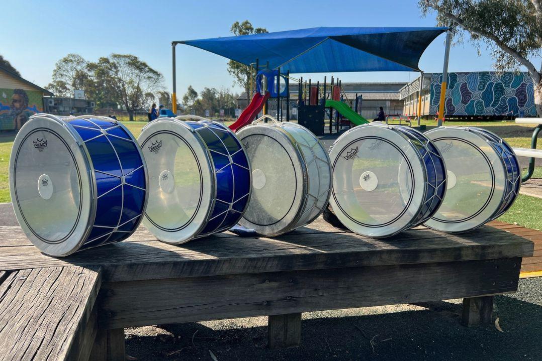 A row of large blue-and-silver drums sits on a wooden platform in a sunny playground, with a shade canopy, slides, and mural-covered walls behind them.