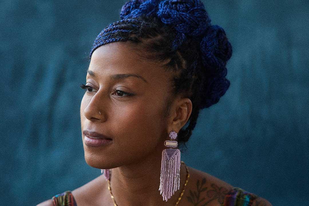 A thoughtful Black woman with vivid blue braided hair, long lavender earrings, and a patterned top gazes to the side against a textured teal backdrop.