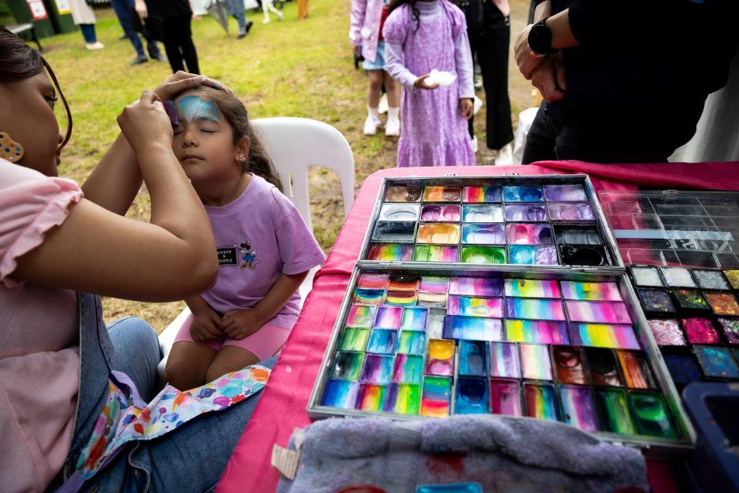 At an outdoor fair, a woman gently paints a young girl’s face blue beside a colorful face-paint palette, creating a calm, cheerful scene.