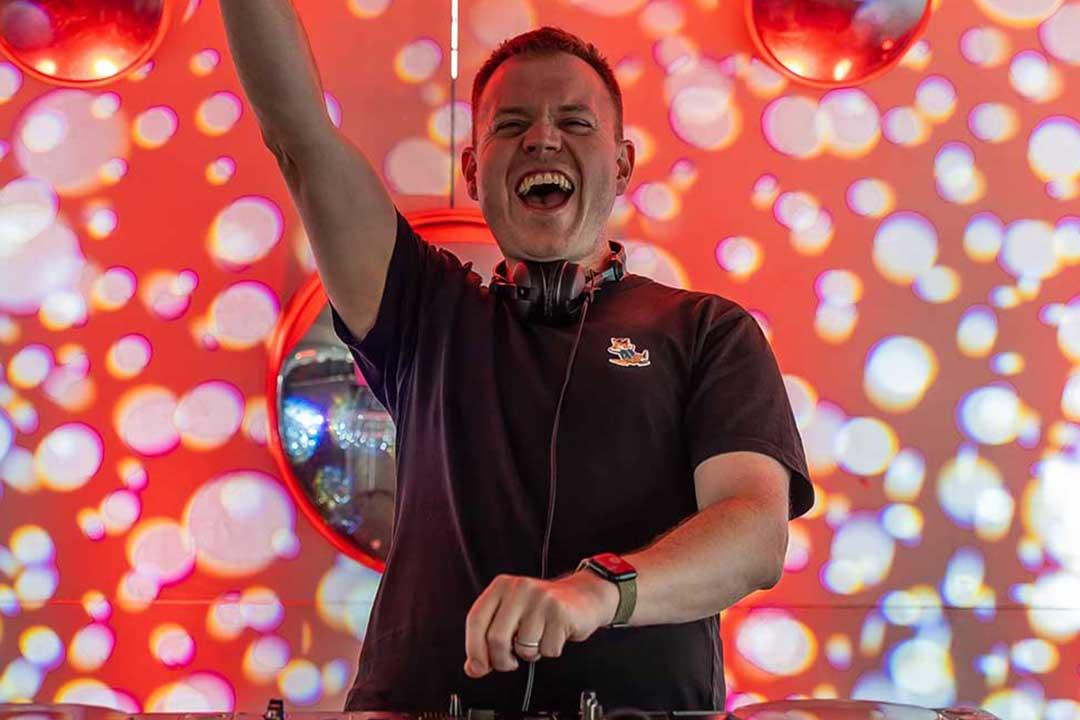 A jubilant DJ raises one arm behind the decks, headphones around his neck, amid vivid red and pink lights and glowing bokeh at a lively music event.