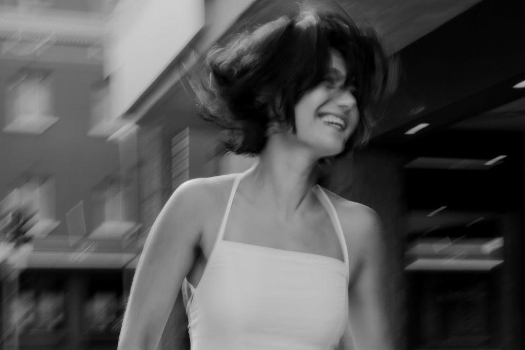Black-and-white, motion-blurred photo of a woman in a white halter top laughing as she turns, her short hair swinging; the mood is lively and carefree.
