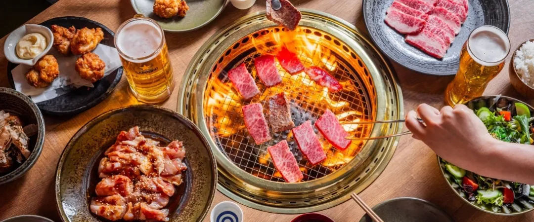 Table with grill in the middle and hands putting raw meat to cook. 