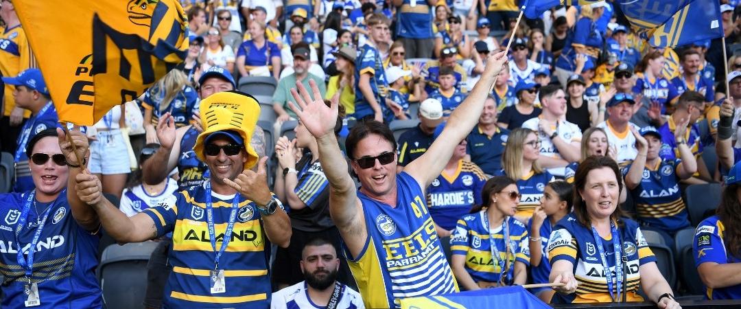 A crowd of enthusiastic sports fans in blue and yellow jerseys cheer at a stadium, waving flags and banners. The atmosphere is vibrant and joyous.