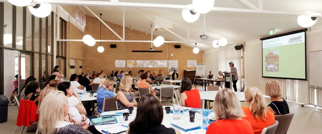 Large room with multiple round with 10 people sitting at each looking at a presenter at power point screen