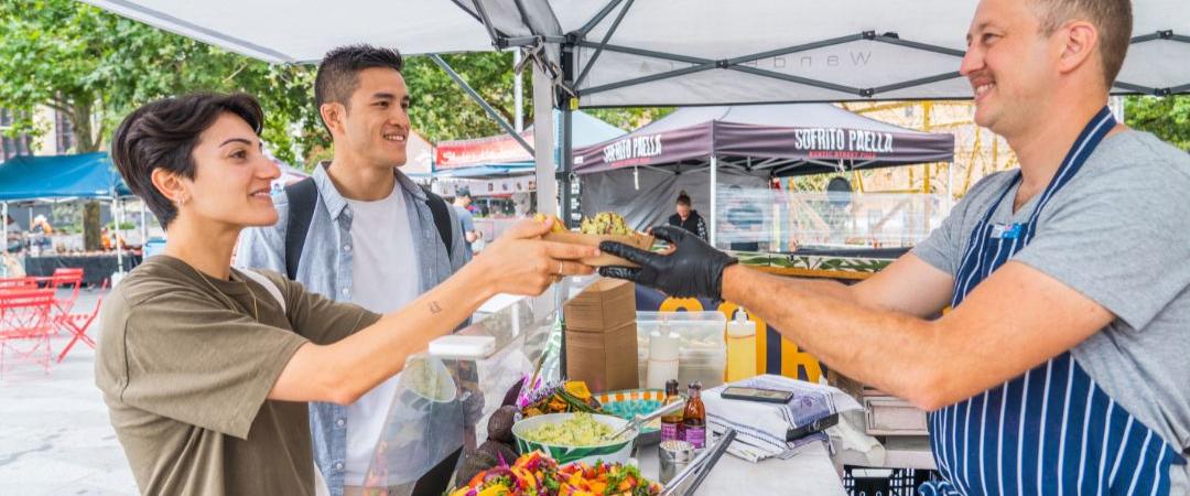 Two people smiling and receiving food from a vendor at an outdoor market stall. Fresh produce is displayed, and trees are visible in the background.