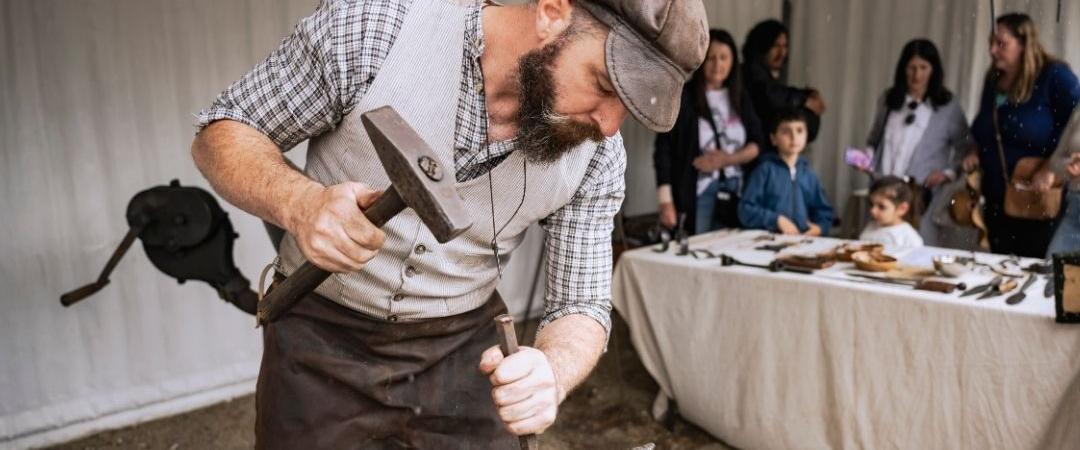 A blacksmith in a hat and apron intensely works with tools on a piece of metal. Onlookers watch from a distance in a workshop setting.