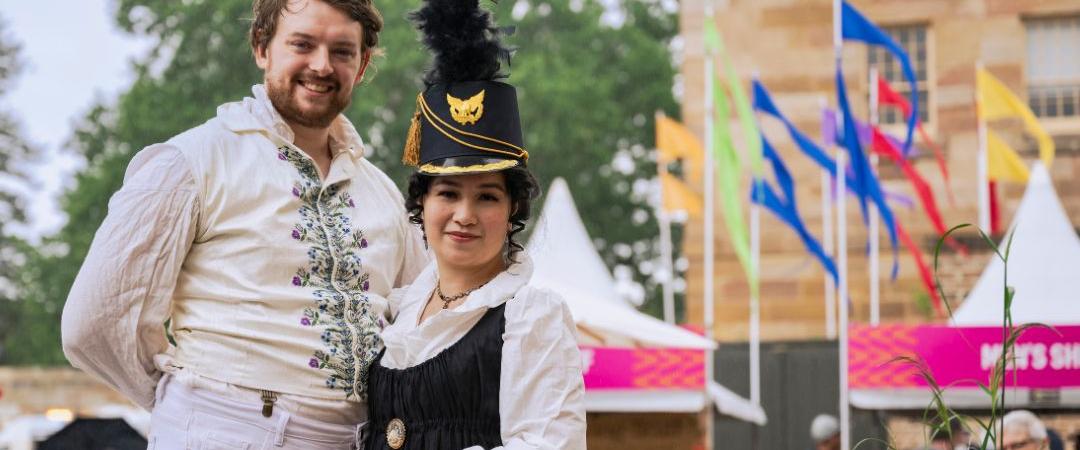 Two people in historical costumes pose cheerfully outside a building with flags. The scene conveys a festive and celebratory atmosphere.