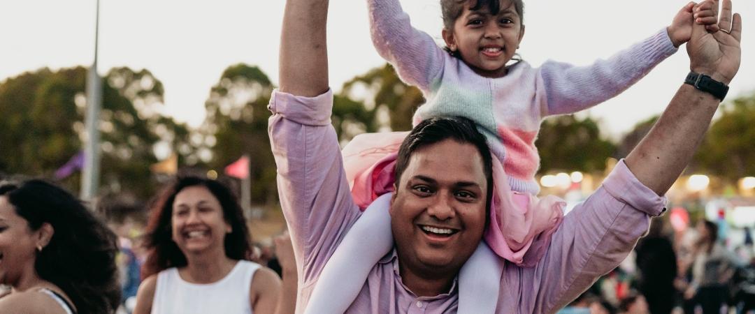 A cheerful man, in a pink shirt, holds a smiling young girl on his shoulders at an outdoor event. People and trees in the background suggest a lively, joyful scene.