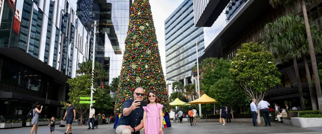 A man and girl take a selfie near a large, colorful Christmas tree in a modern city plaza, surrounded by tall buildings and casual onlookers.