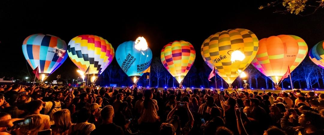 A row of colourful hot air balloons glow at night, illuminating a large crowd of spectators. The scene conveys excitement and a festive atmosphere.