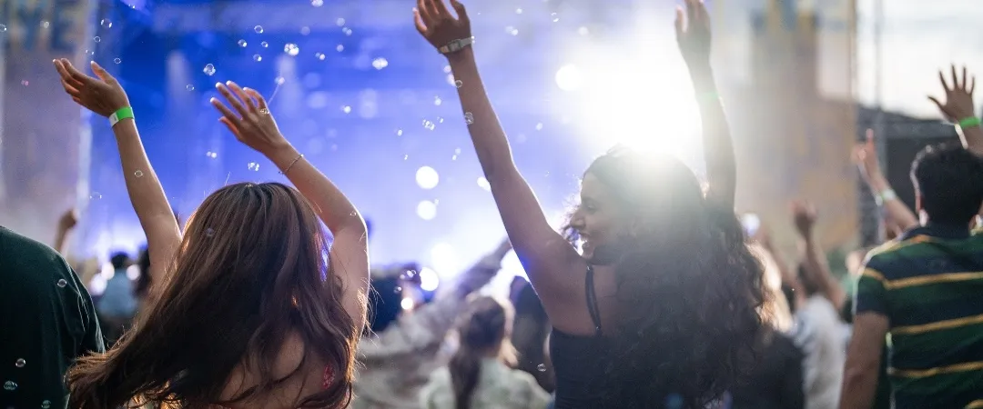 Concert crowd with raised hands enjoys live music under bright stage lights. The atmosphere is energetic and lively, capturing a sense of excitement.