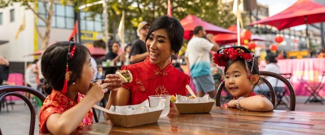 A woman in a red shirt with two children sits at an outdoor table enjoying ice cream. The children smile, wearing red and Minnie Mouse ears. Bright, cheerful atmosphere.