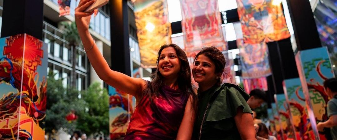 Two people take a joyful selfie amidst vibrant city billboards featuring colourful graphics and advertisements, creating a lively, urban atmosphere.