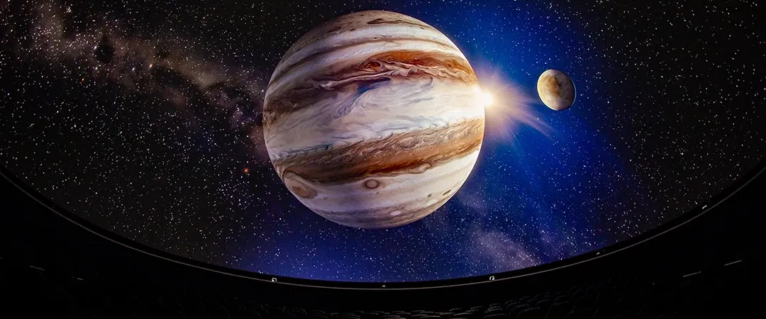 A planetarium display shows a vivid image of Jupiter and its moon against a starry backdrop. Empty theater seats in the foreground add to the immersive feel.