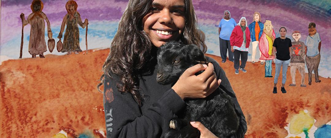 An indigenous girl holding a goat with a bright artwork behind her