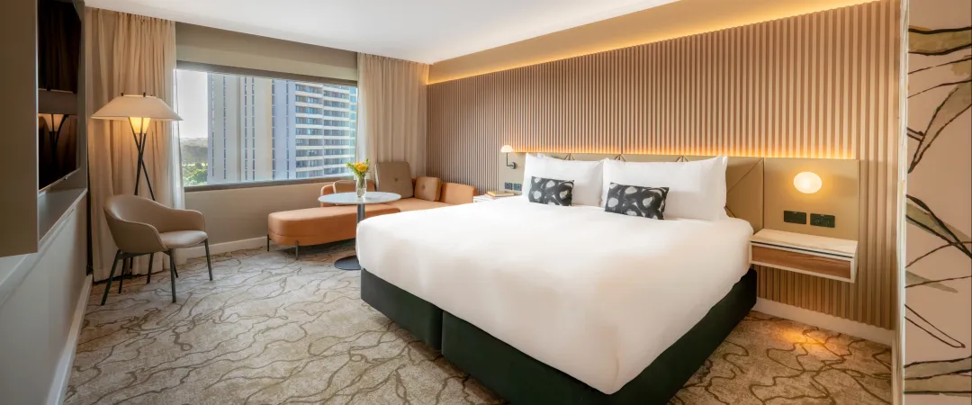 Modern hotel room with a large, white bed, abstract carpet, and a city view. Warm lighting and stylish decor create a comfortable, inviting atmosphere.