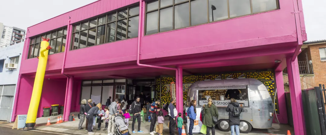 A group of people stands outside a pink building next to a silver truck parked on the street.