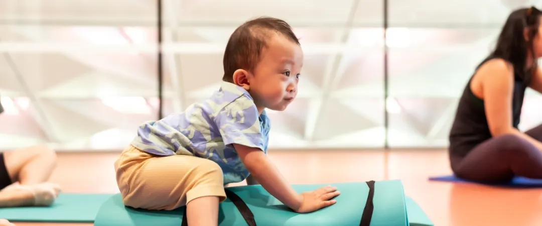 A toddler in a patterned shirt and beige pants balances on a yoga mat indoors, with adults nearby. The scene is lively, playful, and engaging.