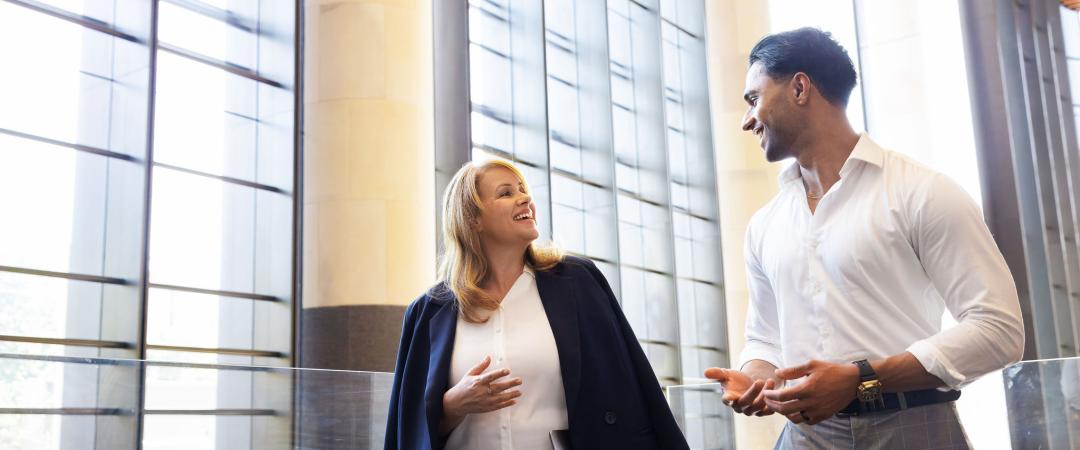 A man and a woman in business attire walk and converse in a bright, modern atrium.