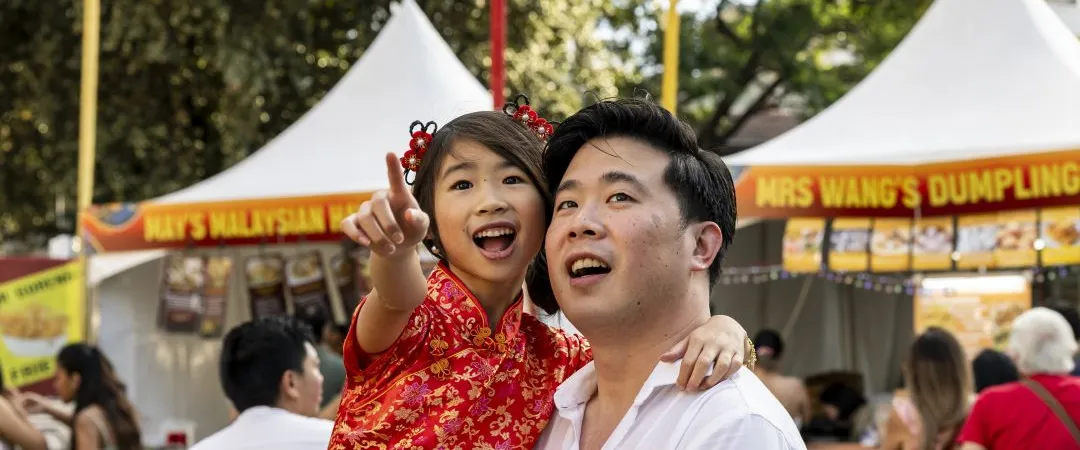 At a lively festival, a smiling father holds a young girl in a red Chinese dress as she points excitedly near Malaysian dumpling stalls.