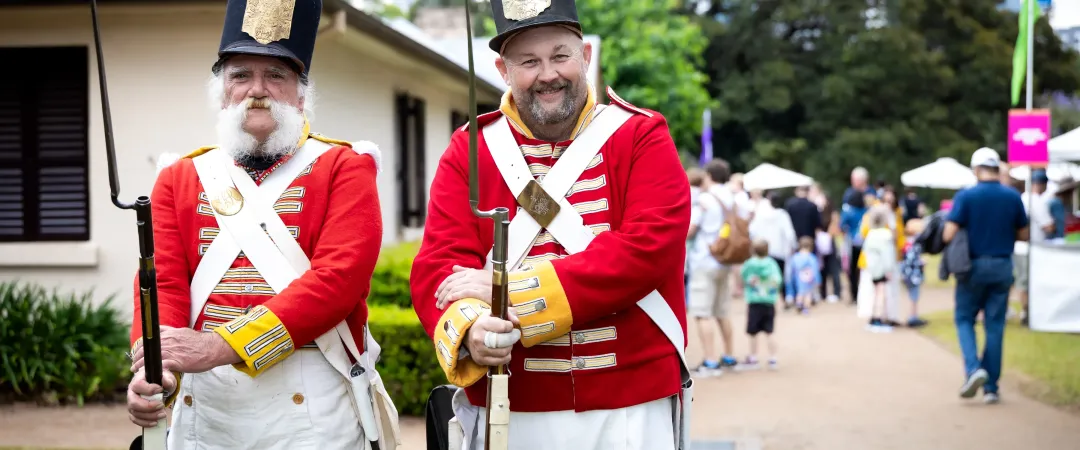 Two historical reenactors dressed in bright red military uniforms and tall black hats stand outdoors holding replica muskets.