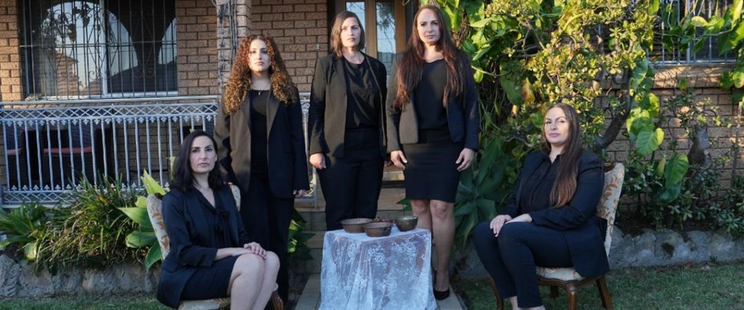 Five women in black clothing pose solemnly in front of a brick house, with leafy greenery as a backdrop.