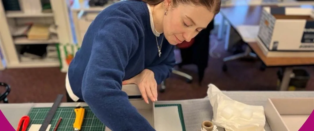 Focused woman in a blue sweater assembles packaging at a craft table, surrounded by cutting tools, cardboard, tissue paper, and studio supplies.