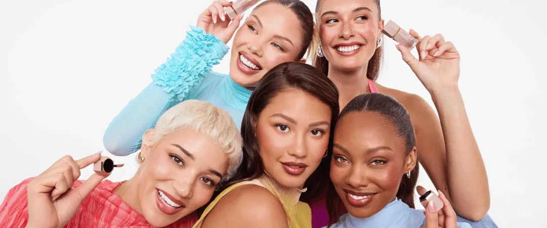 Five women in colorful outfits smiling and holding beauty products against a white background. The image conveys joy and camaraderie.