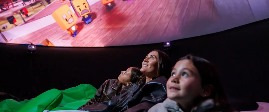 A family of three sits on bean bags, smiling and looking at a dome screen projecting colorful animated square characters, conveying joy and wonder.