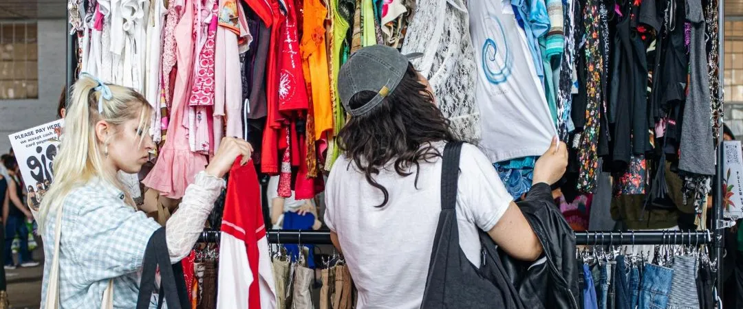 Two people browse colorful clothes on racks at a busy market. They seem focused and engaged, each holding items, conveying a sense of interest and exploration.