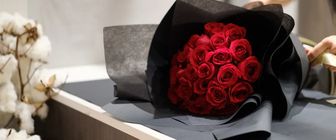 A bouquet of vibrant red roses wrapped in black paper is being held by a person. Cotton buds in the background add a soft, elegant contrast.