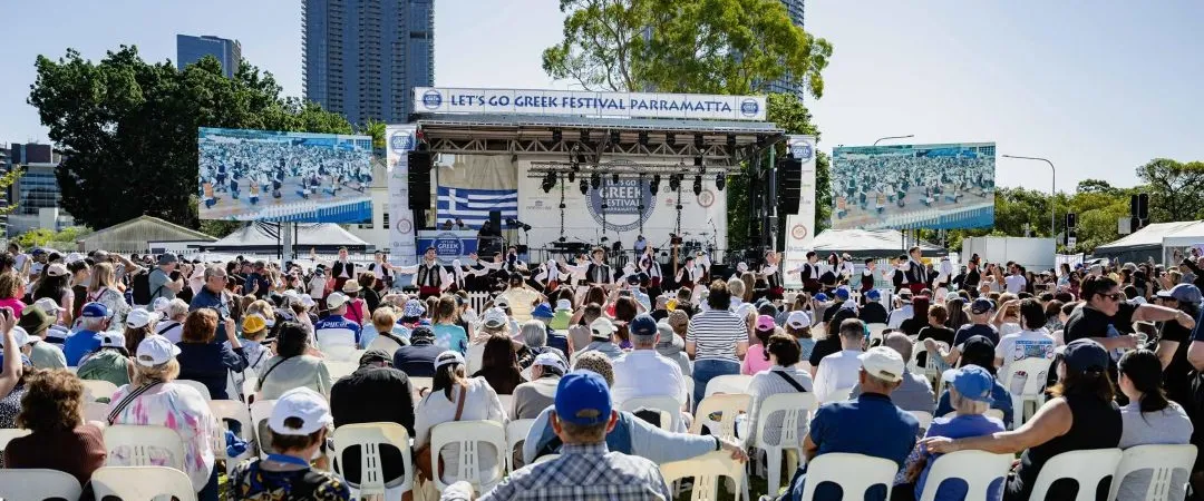 Crowd seated on white chairs at Let's Go Greek Festival Parramatta, watching a lively outdoor stage with skyscrapers behind it.