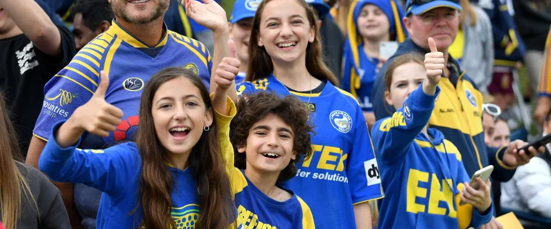 A group of enthusiastic fans wearing blue and yellow jerseys, some giving thumbs up and smiling, in a lively crowd at a sports event.