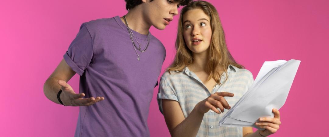 A young man and woman discuss a document against a vibrant pink background. The woman points at the paper, engaging the surprised man. Casual and focused mood.