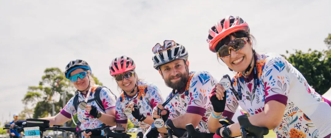 Four cyclists in floral jerseys and helmets smile and hold medals next to their bikes on a sunny day. The scene is cheerful and celebratory.