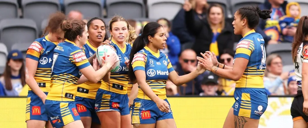 Women’s rugby players in blue-and-yellow uniforms huddle and celebrate on the field, while teammates clap and fans watch behind them.