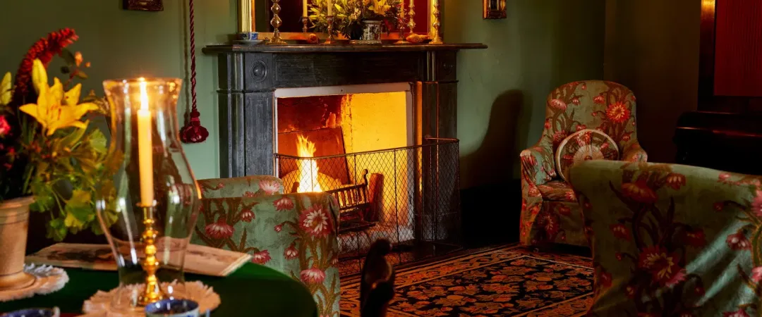 A warmly lit vintage living room with a glowing fireplace, candlelit mantel, floral armchairs, tea service, flowers, and a curious black cat.