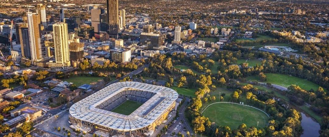Aerial view of a cityscape at dusk with a brightly lit stadium in the foreground. Skyscrapers and green park areas surround the stadium. The sky is blue with clouds.